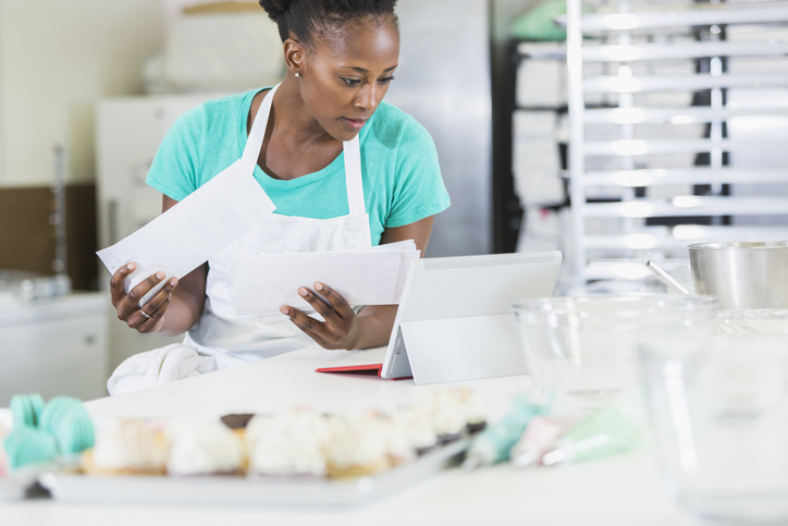 A small business owner of a bakery, paying bills on her laptop computer. She is in a commercial kitchen surrounded by baked goods and equipment, wearing an apron, looking down at the screen.